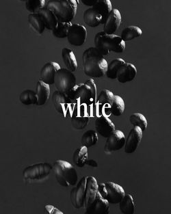 Close-up of dark roasted coffee beans falling against a black background with the word 'white' overlayed in the center.