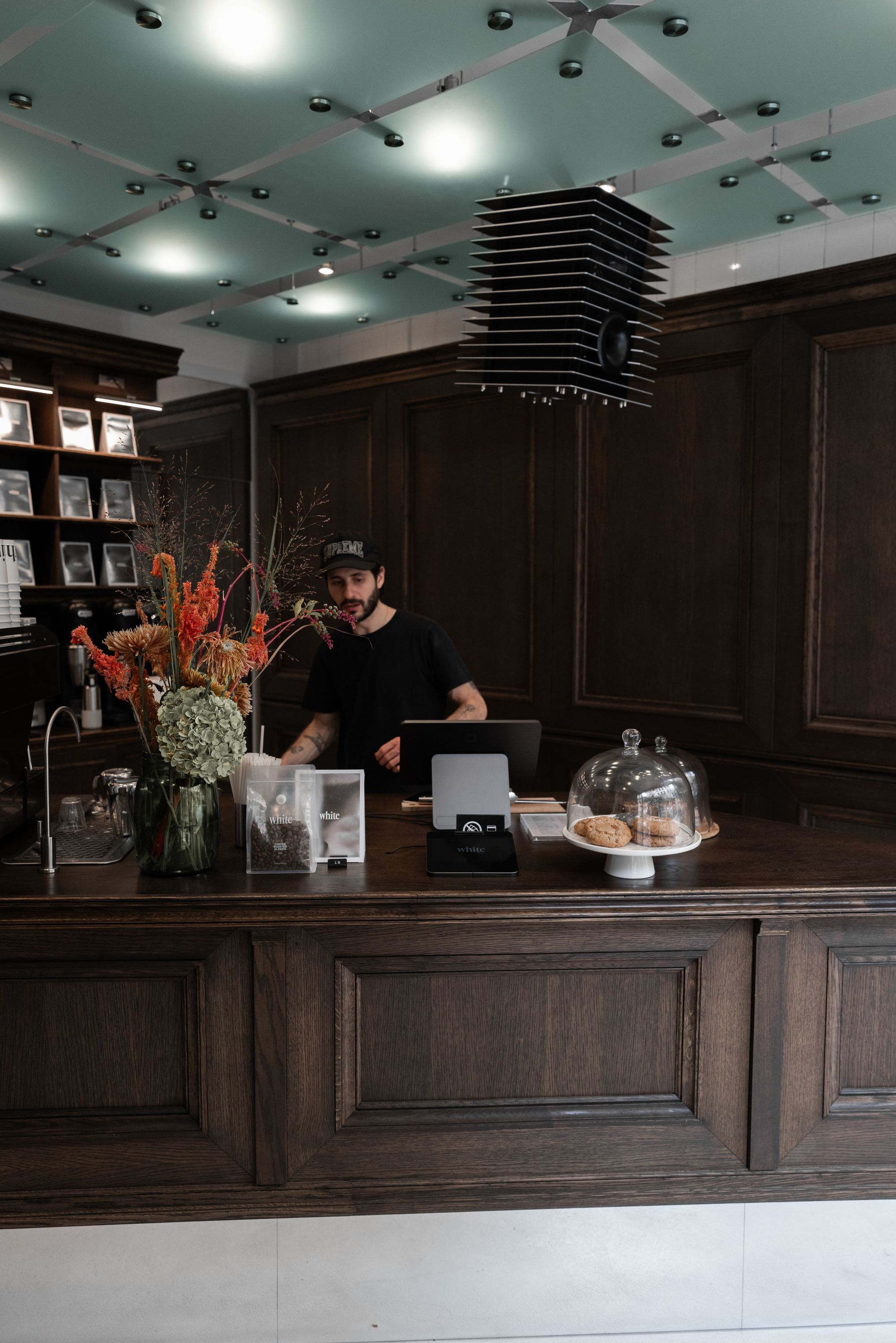 Dark wood counter at a coffee shop with a barista working behind it, flowers, and pastries on display.
