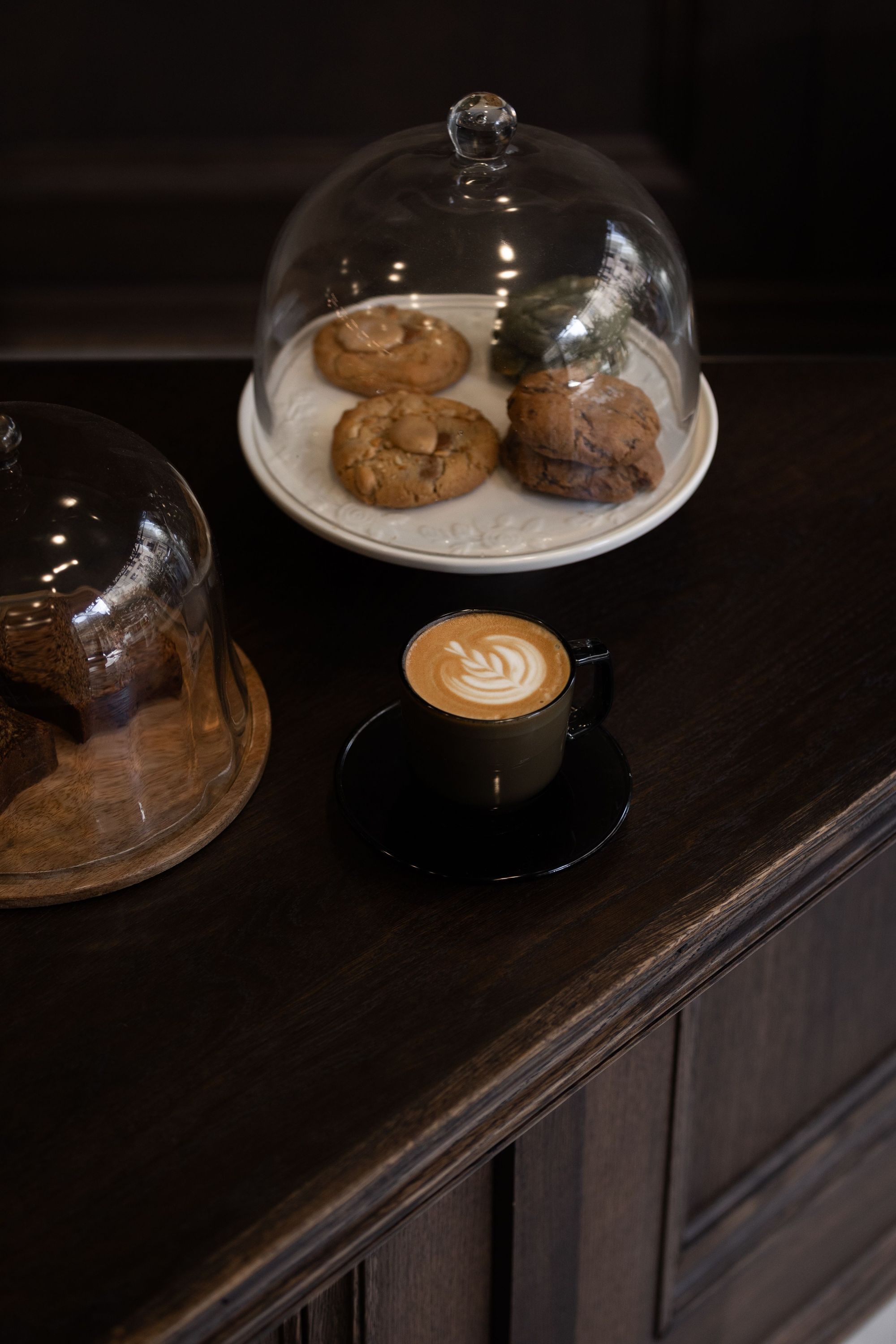 Tasse de café avec latte art accompagnée de biscuits sous cloche sur un comptoir en bois foncé.