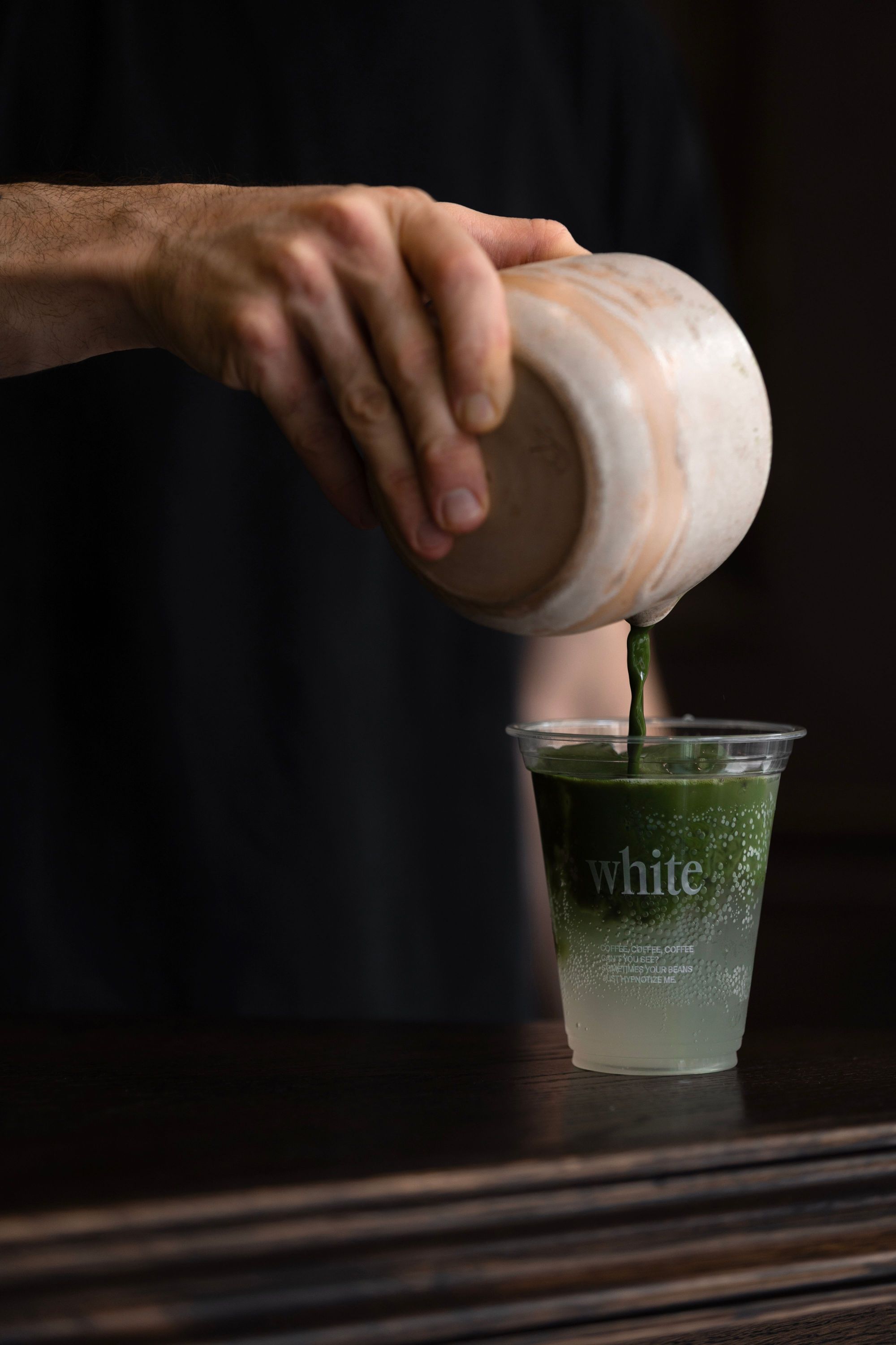 Un verre transparent avec logo white où un liquide vert est versé à partir d'un récipient près d'une main sur un fond sombre.